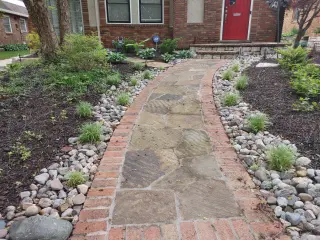 Brick-bordered flagstone front walkway leading to a red door, lined with river rock and new plantings